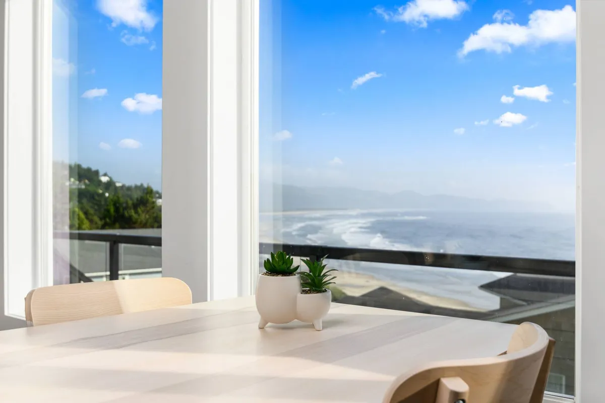 This image features a serene dining area set against large windows that frame a panoramic view of the ocean. The space is accented with potted plants and a four-person dining table, emphasizing both the connection to nature and the opportunity for shared meals or quiet reflection.