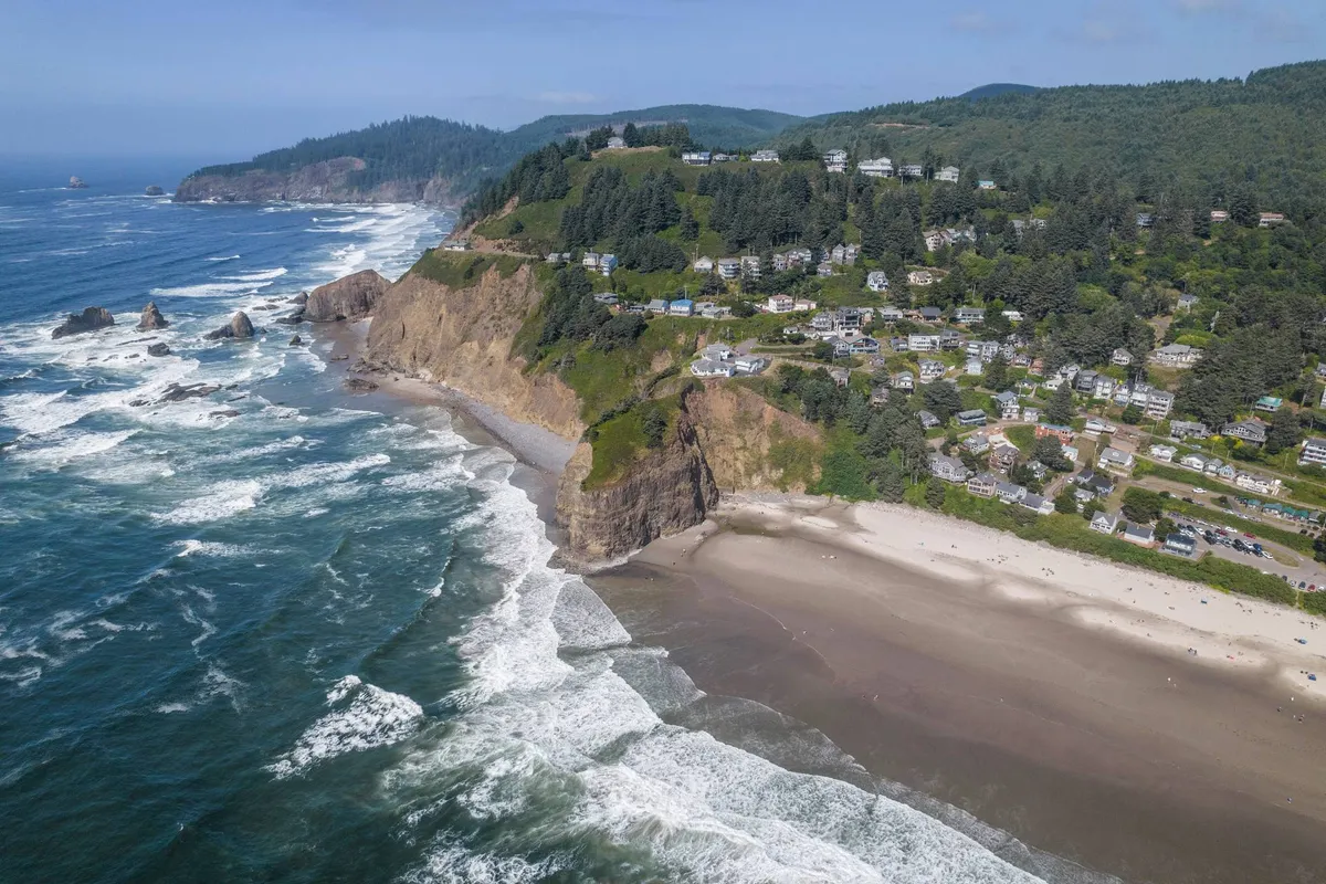 This image displays the scenic cliffside homes of Oceanside, the expansive beach below, rolling ocean waves, and a lush forest backdrop. The scene is tranquil and showcases the natural beauty and pet-friendly beach environment just steps away from Vista House.
