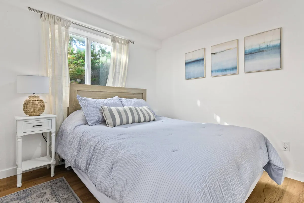 A tastefully decorated bedroom with a comfortable bed, soft textiles, calming artwork above the headboard, a lamp on the nightstand, and a large window that lets in gentle natural light. The setting appears tranquil and perfect for relaxation, with subtle coastal touches in the decor.