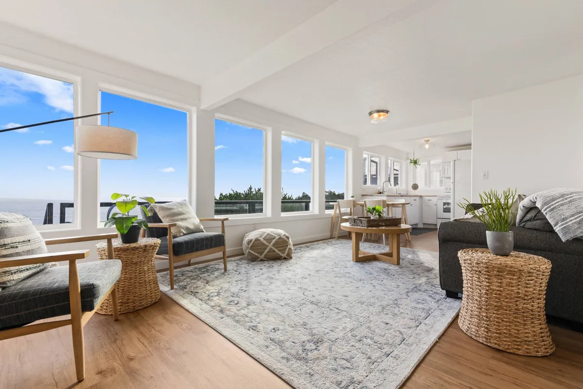 The image showcases a tranquil living area with cozy seating arranged around a round wooden coffee table, a textured area rug, and expansive windows framing breathtaking panoramic views of the Pacific Ocean. Soft natural light pours in, highlighting the inviting space and the seamless connection to the coastal landscape.