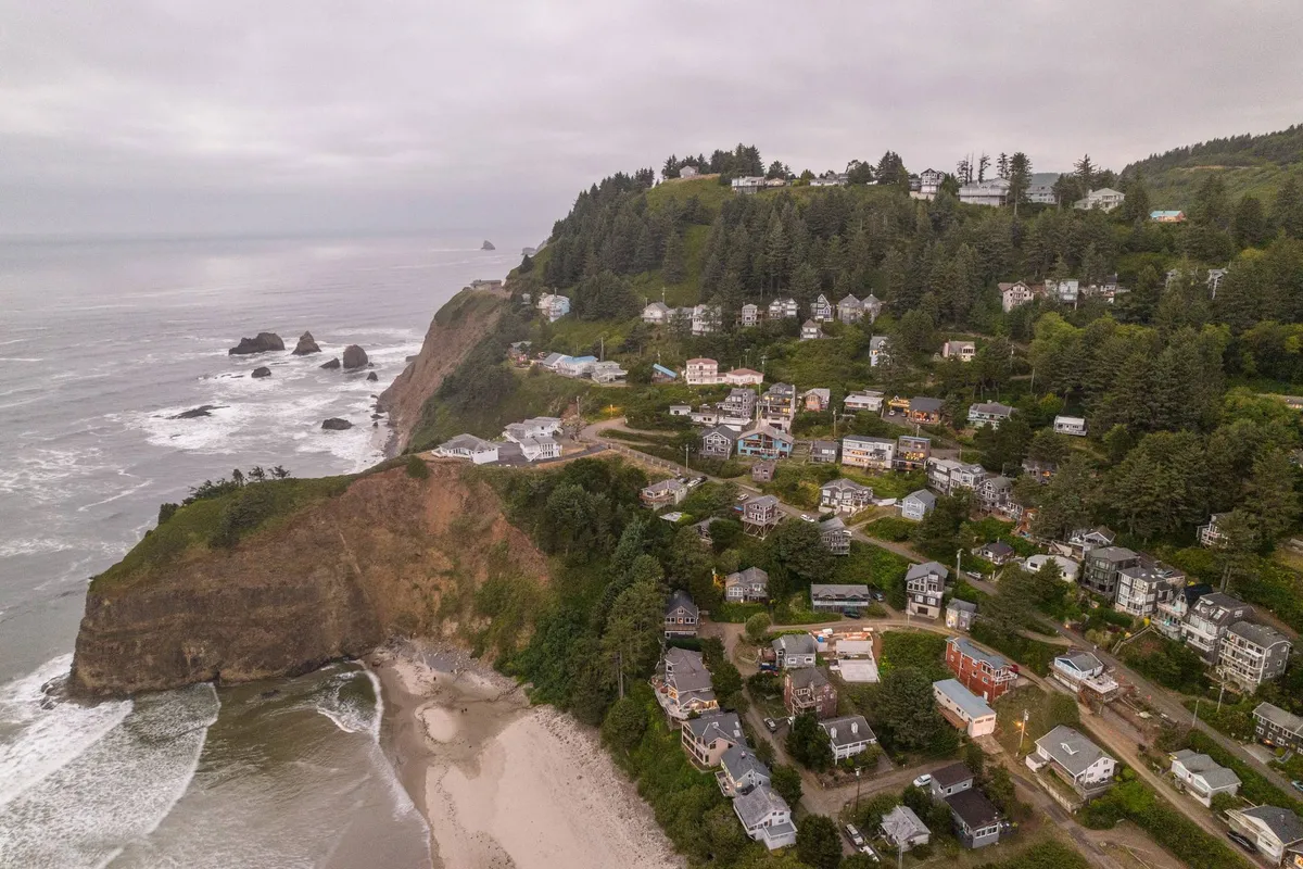 A panoramic view from the cliffs, capturing the dramatic coastline, sandy beach, and the Pacific Ocean stretching into the horizon. Houses perched nearby suggest a community feel, while the setting provides a natural, immersive connection to the outside environment.