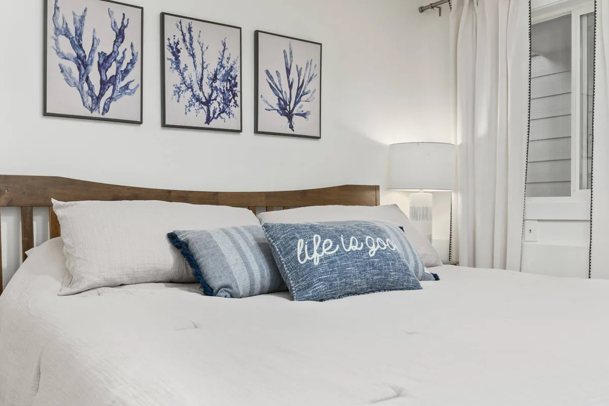A welcoming bedroom with decorative pillows, framed coral artwork nodding to the coastal theme, and a lamp for cozy reading sessions. The bed looks inviting, and the overall mood is serene and inviting.