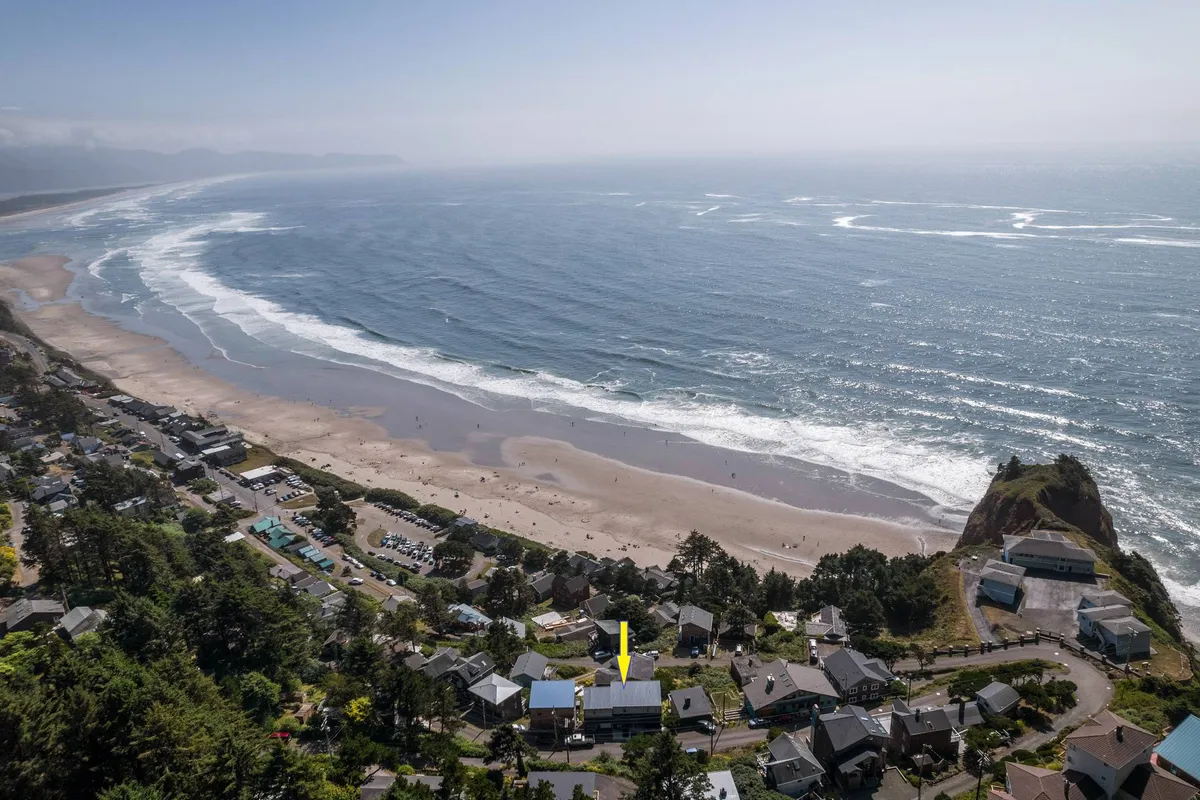 A wide-angle view captures the ocean, sandy beach, and a scattering of homes along the shoreline, with a picturesque rocky outcrop in the distance. The scene is tranquil, ideal for showcasing sunrise or sunset walks, and the mood invites guests to relax and take in the beauty of the coastline.