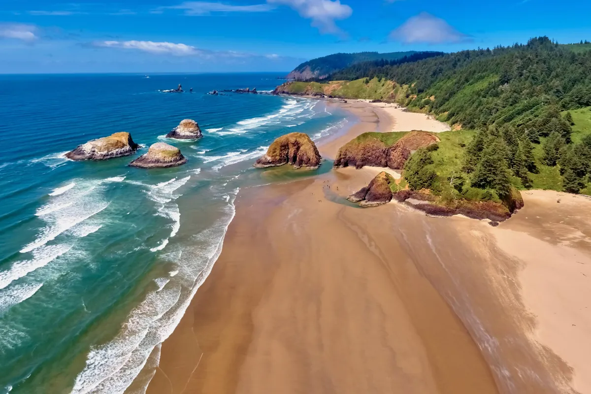 A tranquil coastal landscape with striking rock formations along the shore, gentle waves breaking on the sand, and lush, forested hills in the background. The golden light suggests sunset, adding to the sense of peace and natural splendor.