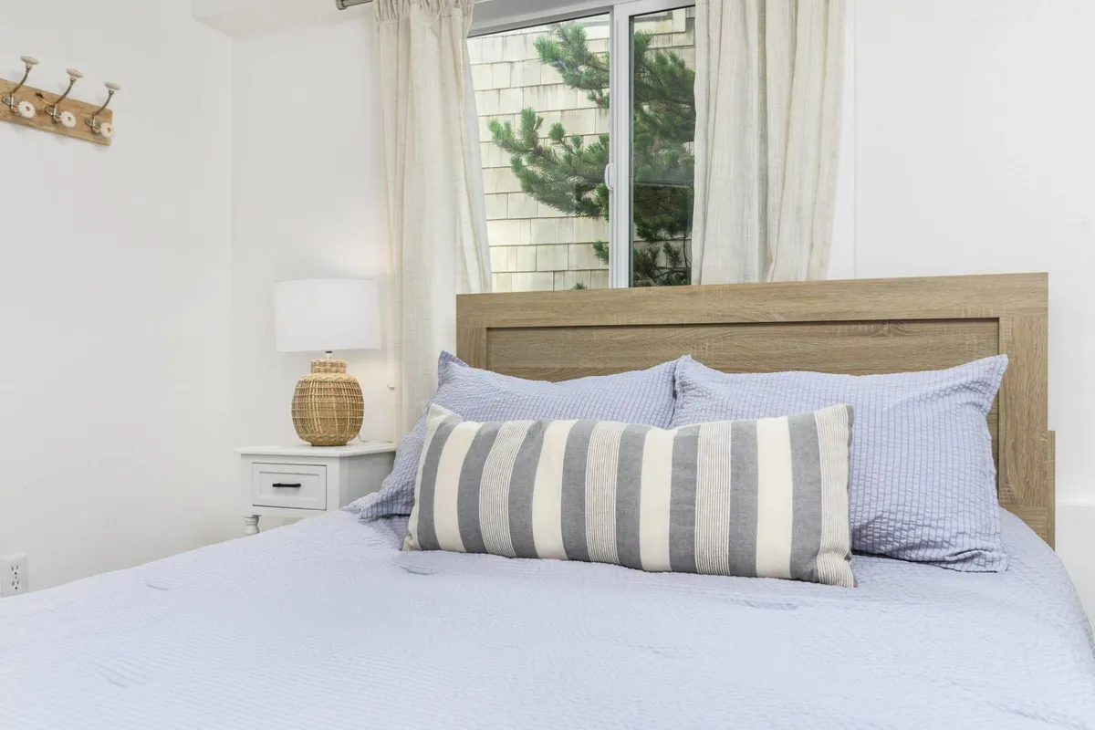 A cozy bedroom scene featuring a bed adorned with a striped pillow and crisp linens, a bedside table with a lamp, and a window offering a view of lush greenery outside. The light filtering through the window creates a serene atmosphere.