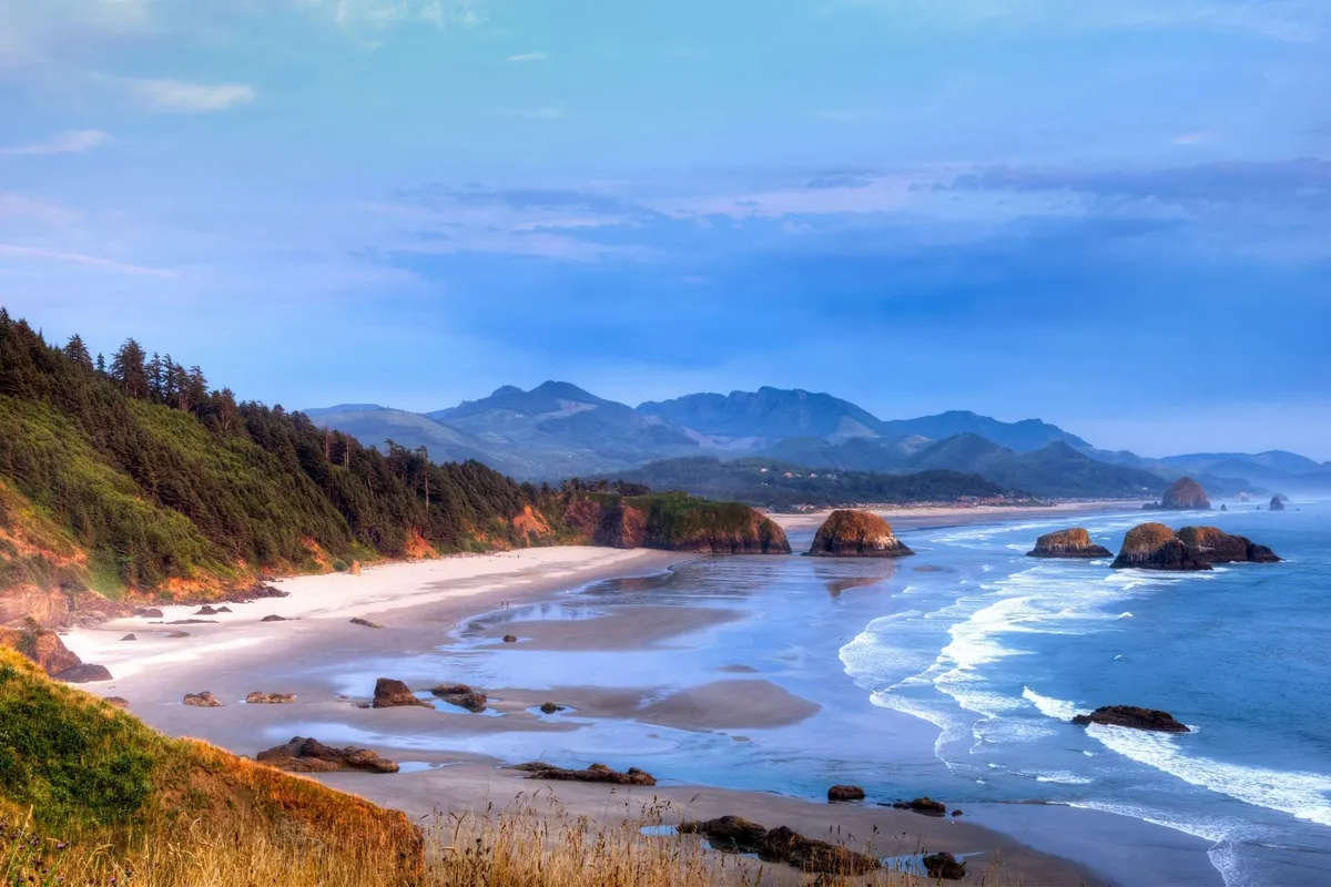 A sweeping view of the Oregon coast featuring a broad sandy beach, dramatic rock formations jutting from the surf, and distant mountains rising beyond the shoreline. The scene is peaceful, with soft light and a serene atmosphere that evokes the natural beauty and grandeur of the area.