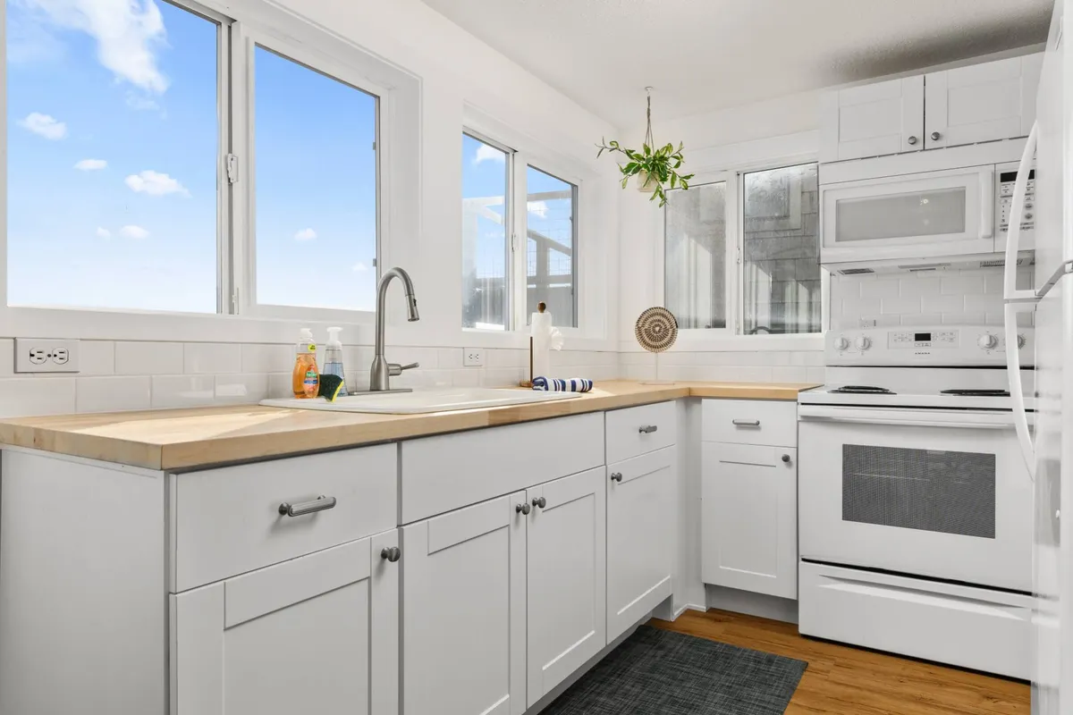 A bright, cheerful kitchen corner with a modern sink, wooden countertops, a large window bringing in morning light, and a green hanging plant. The setting feels fresh and inviting, perfect for sharing a coffee or preparing meals.