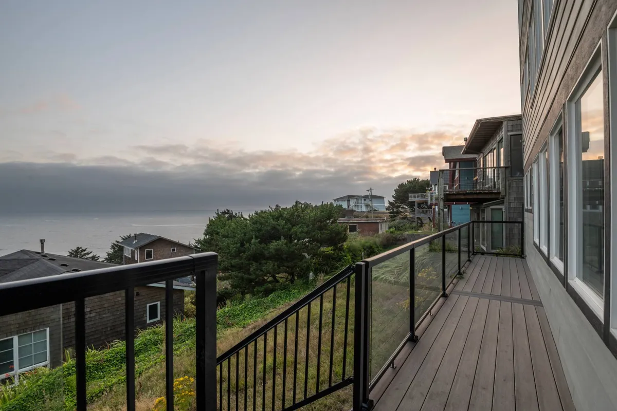 A serene exterior view featuring a balcony railing with comfortable seating, overlooking the expansive ocean. The sun is setting, casting a warm glow over the water and lush greenery surrounds the space, offering a tranquil environment perfect for relaxing outdoors.