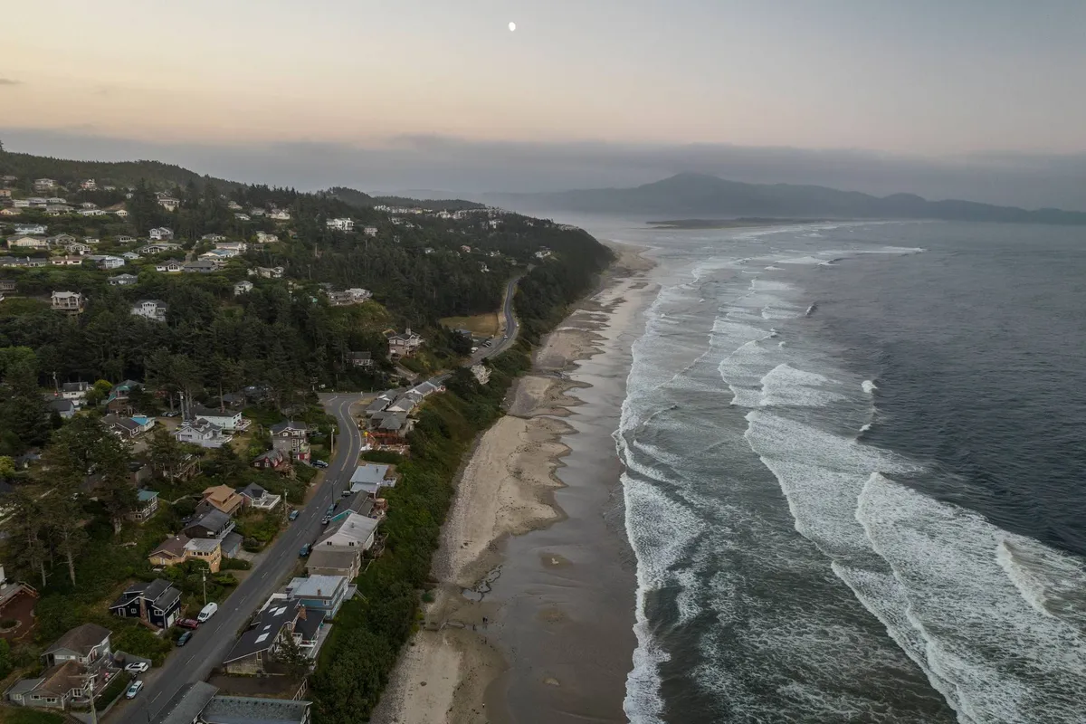 A scenic overlook from a hillside showing ocean waves crashing along a coastal road, hillside homes, and the soft glow of the moon above. The view blends natural and human elements, emphasizing the proximity to both wild landscapes and comfortable retreats.